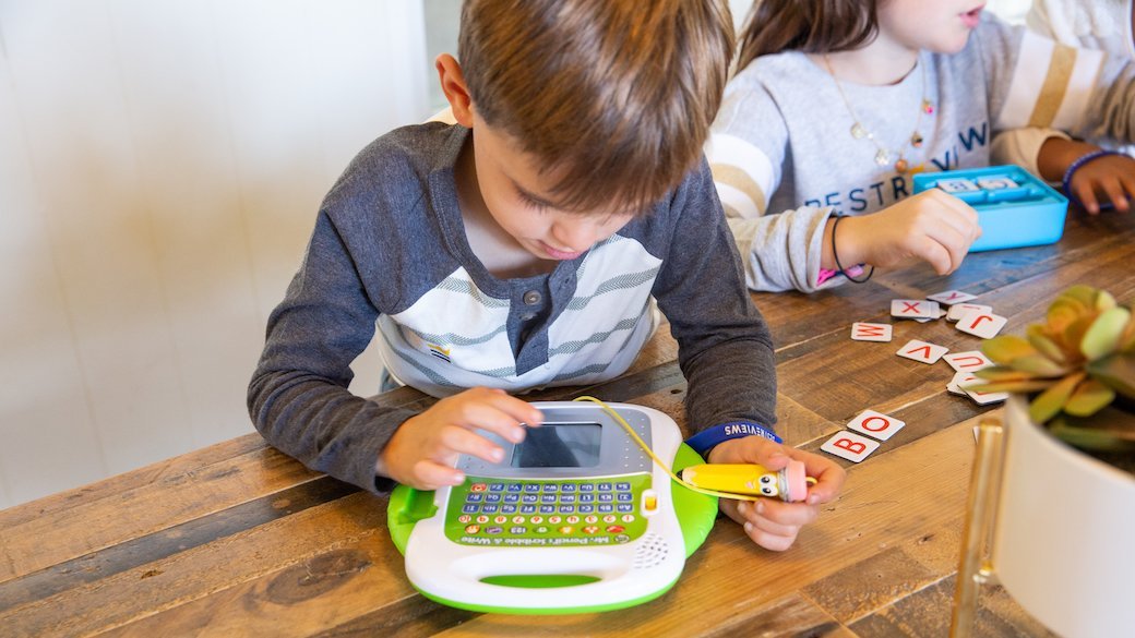 A short haired child wearing a gray and white long-sleeved shirt kneels at a table and plays with a white and bright green interactive toy. In the background, a long haired child in a light gray shirt plays with a blue toy.